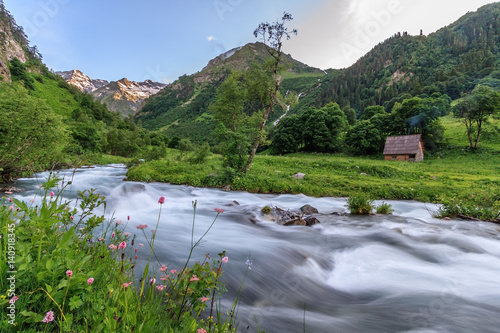 Beautiful scenic landscape of rapid Imeretinka mountain river valley with snowy mountain peaks in Caucasus mountains at late spring. Wooden cottage in the forest by the river bank