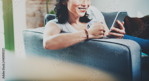 Young smiling Asian woman spending rest time at home on sofa, holding ceramic cup at hand and using smartphone for video conversation with friends.Blurred background, crop.Wide. photo