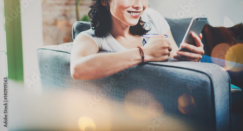 Young smiling Asian woman spending rest time at home on sofa, holding ceramic cup at hand and using smartphone for video conversation with friends.Blurred background, flares effect.Wide. photo