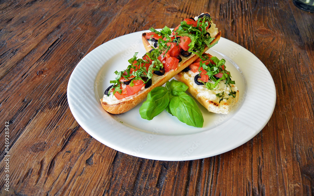 Preparing delicious Italian tomato bruschetta with chopped vegetables, olives, cheese and oil on grilled or toasted crusty baguette sprinkled with seasoning and spices on a plate on wooden background Preparing delicious Italian tomato bruschetta with chopped vegetables, olives, cheese and oil on grilled or toasted crusty baguette sprinkled with seasoning and spices on a plate on wooden background