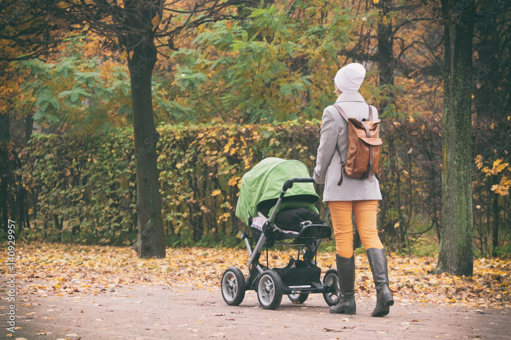 Young mother pushing stroller in autumn park. Young woman strolling with baby in green buggy outdoors Young mother pushing stroller in autumn park. Young woman strolling with baby in green buggy outdoors
