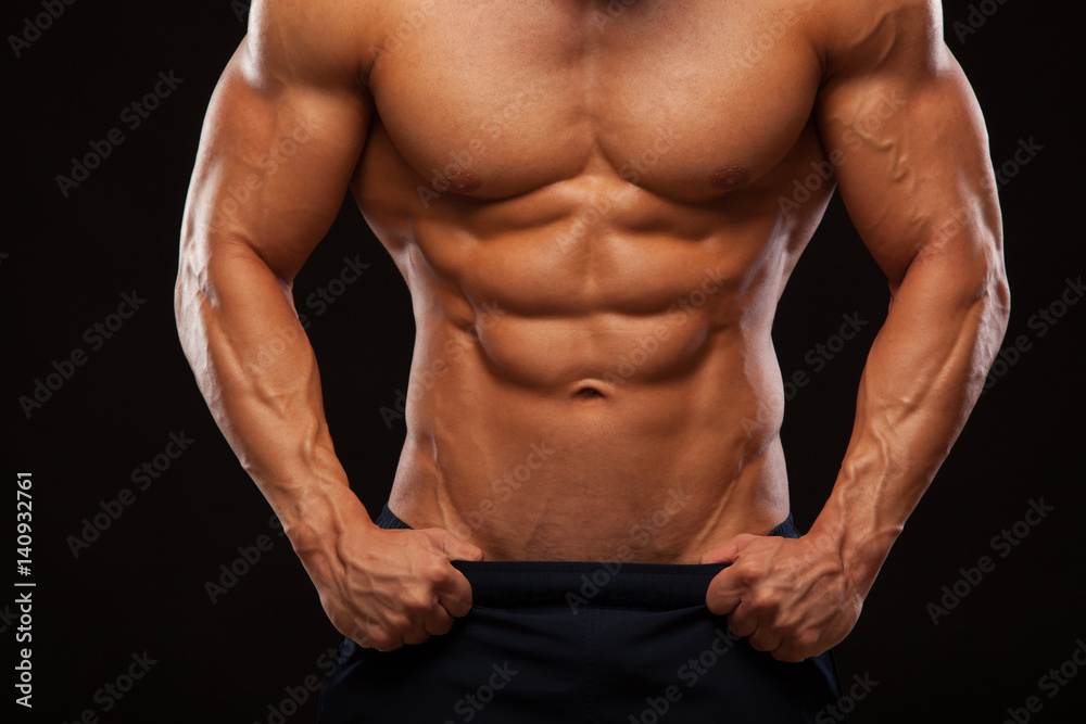 Strong Athletic Man - Fitness Model showing Torso with six pack abs. stands straight and puts his hands in trousers. isolated on black background with copyspace