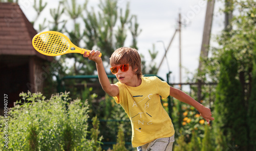 Cute boy wearing orange sunglasses plays beach tennis in the garden