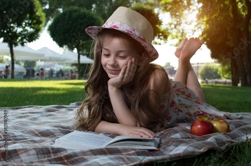 Cute little girl in the wicker hat and dress with flowers reading a book on a sunny summer day lying on the lawn. On open air. training. Reading. Development