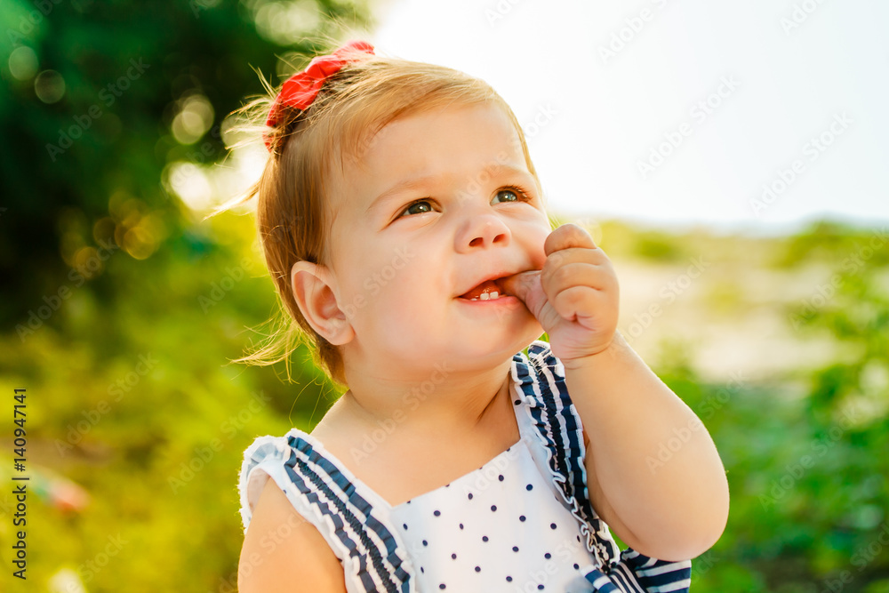 The little girl with short hair suck finger. The baby is looking into the sky. Little beautiful child sitting on the bank of the river among the green trees. The little girl with short hair suck finger. The baby is looking into the sky. Little beautiful child sitting on the bank of the river among the green trees.
