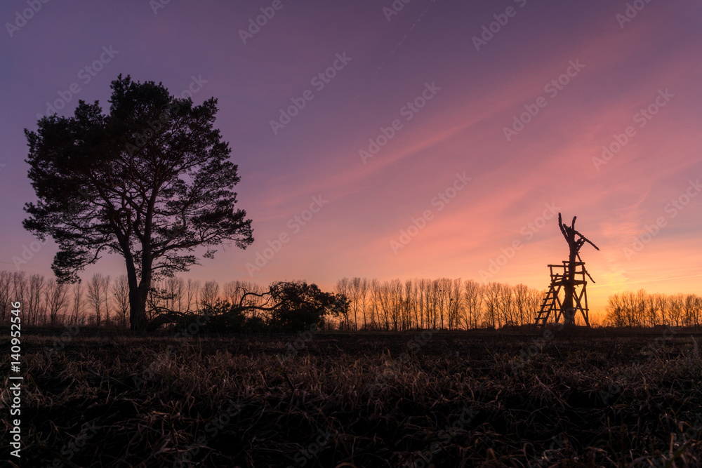 Sonnenuntergang mit Hochstand und Bäumen auf einem Feld Sonnenuntergang mit Hochstand und Bäumen auf einem Feld