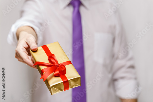 Male hand holding a gift box. Present wrapped with ribbon and bow. Christmas or birthday package. Man in white shirt and necktie. © tanyastock