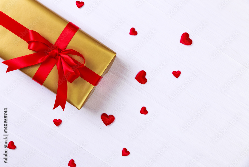 Gift box with red satin hearts. Present wrapped with ribbon and bow. Christmas or birthday golden paper package. On white wooden table. Gift box with red satin hearts. Present wrapped with ribbon and bow. Christmas or birthday golden paper package. On white wooden table.