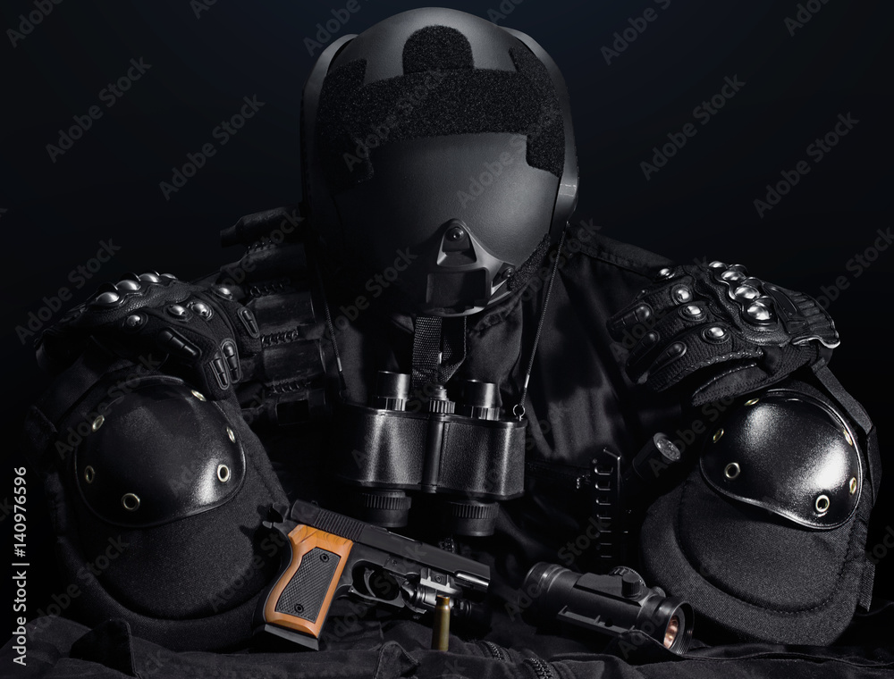 Black military tactical gun, helmet, gloves, cartridge belt, bandolier, gun shell, knife, binocular and knee protection laying on a black table.