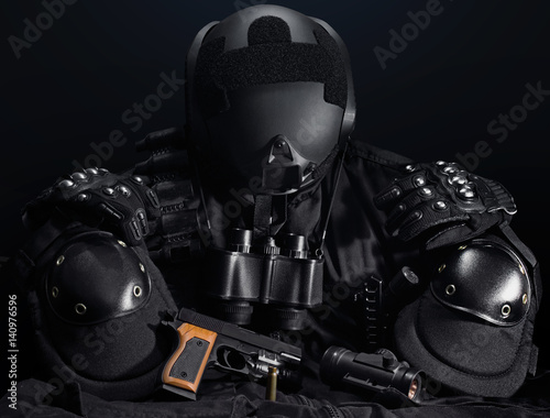 Black military tactical gun, helmet, gloves, cartridge belt, bandolier, gun shell, knife, binocular and knee protection laying on a black table.