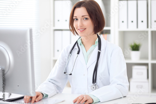 Young brunette female doctor sitting at a desk and working on the computer at the hospital office. Health care, insurance and help concept. Physician ready to examine patient