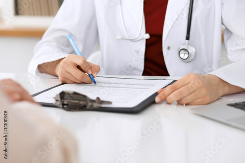 Close up of a female doctor filling up an application form while sitting at the table. Medicine and health care concept