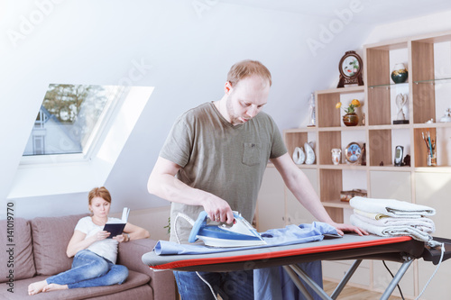 Young woman sitting at the sofa reading the book while her husband is ironing shirt at home.