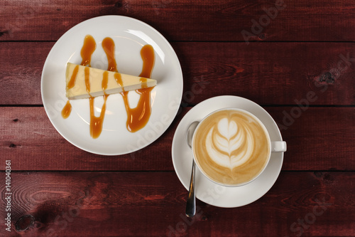 Canvastavla A cup of cappuccino and a piece of cheesecake, drizzled in caramel sauce are in white dish on wooden maroon table
