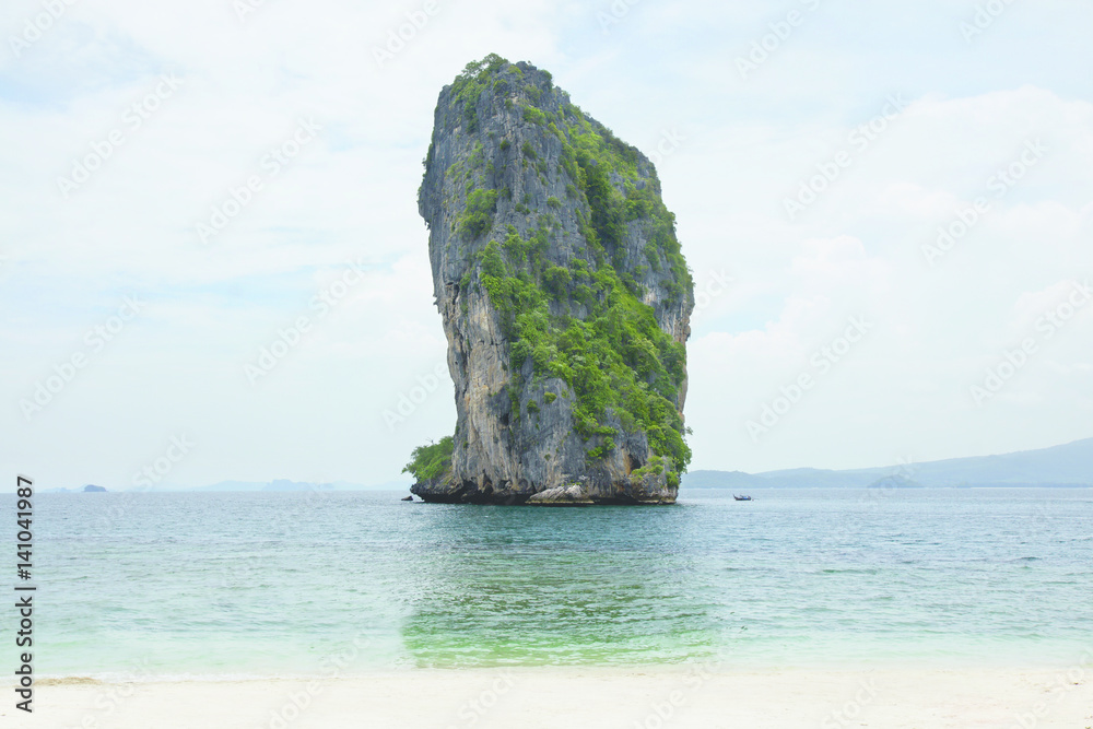 Naklejka premium Big high rock cliff filled with green vegetation surrounded by turquoise blue colored ocean water next to a tropical white sand beach with horizon view at midday, Krabi Thailand.
