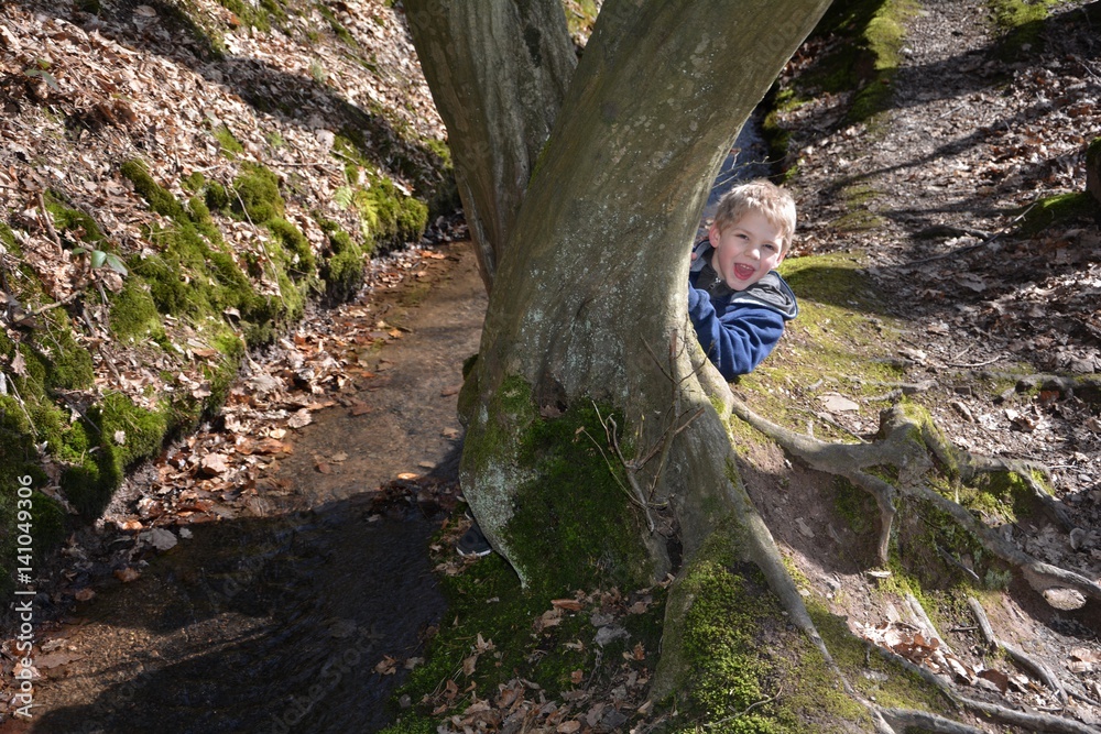 Kleiner Junge  am Bach, kniet hinter einem Baum und lacht  nach vorne