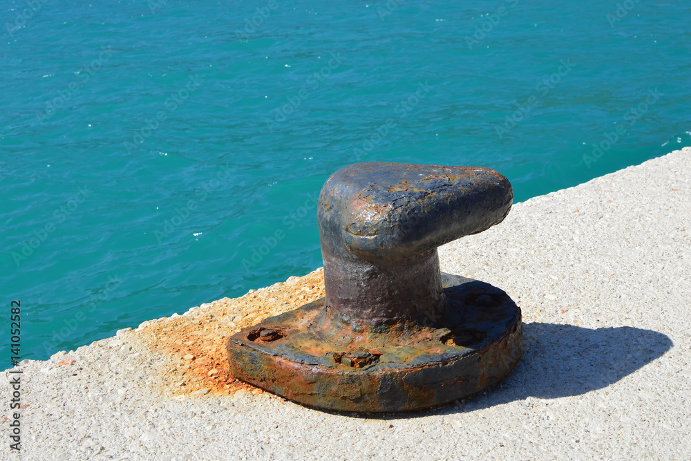 Sunny day with calm blue sea on a pier. Detail of an old rusty bollard Sunny day with calm blue sea on a pier. Detail of an old rusty bollard