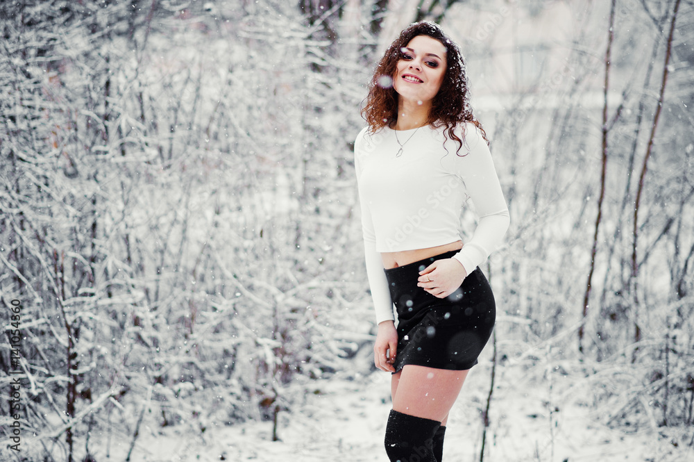 Naklejka premium Curly brunette girl background falling snow, wear on black mini skirt and wool stockings. Model on winter. Fashion portrait at snowy weather. Instagram toned photo.