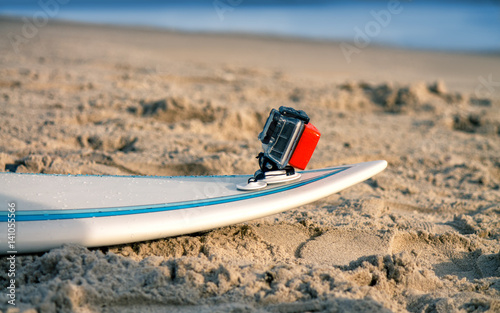 Surfboard with attached action camera lies on the sand on the beach. Action camera is in waterproof case for swimming. photo