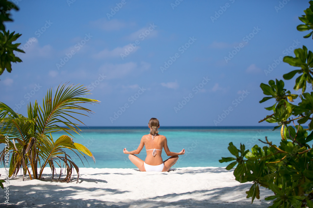 Young woman practicing yoga meditation on the beach facing the ocean near a palm tree on Maldives Young woman practicing yoga meditation on the beach facing the ocean near a palm tree on Maldives