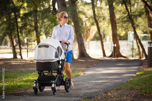 Young mother walking and pushing a stroller in the park. Mother strolling with newborn. Beautiful happy mother with pram outdoors. Summer walks in the sunny day. Girl with carriage. photo