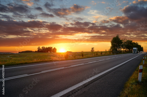 Sunset over the asphalt road in a rural landscape, truck and passenger cars arriving from afar.