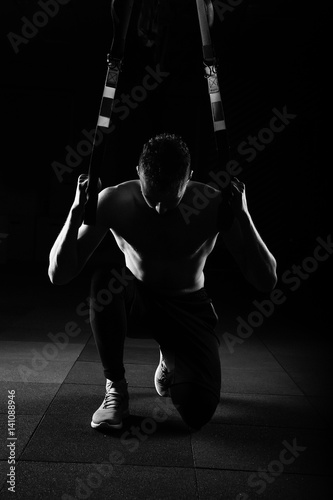 One young handsome brave curly bearded muscled man with a beard makes Total Resistance eXercises fitness trx straps working out in gym crossfit black and white stylised.