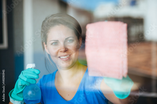 People, housework and housekeeping concept - happy woman in gloves cleaning window with rag and cleanser spray at home