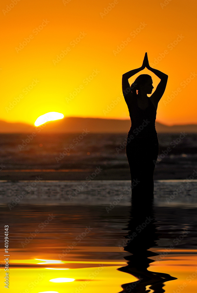 Girl performs yoga pose on beach with warm sunrise on horizon.