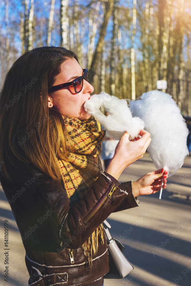 Spring day in the park. Young attractive brunette girl eating sweet cotton wool.