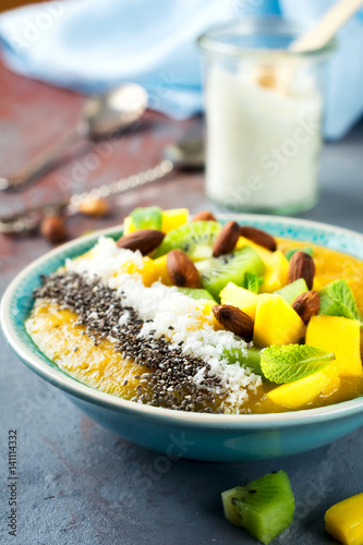 Mango smoothie bowl with pieces of kiwi, mango, coconut shavings and chia seeds on a stone background. Selective focus.