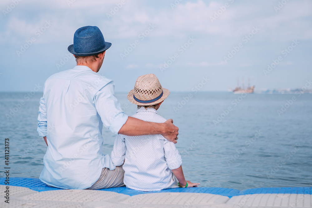 Father and son sit on the sea pier and look on the ship on horizont line