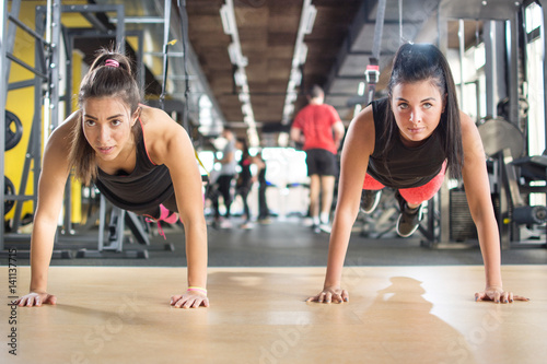 Two sporty girls doing push ups while their legs are on suspension strap at gym.