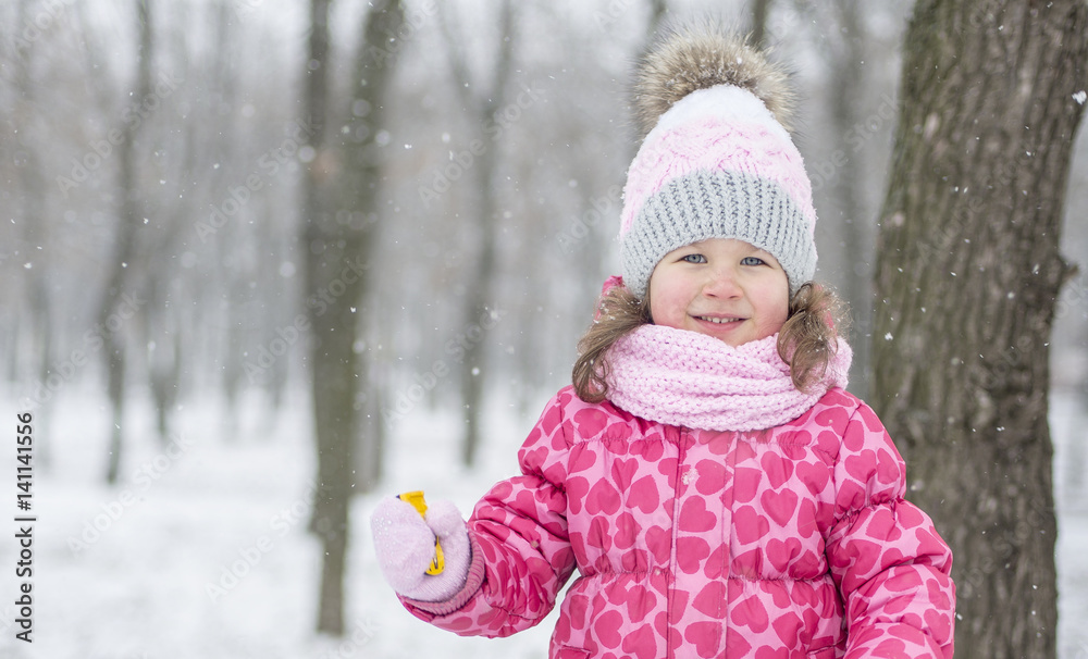 A small beautiful girl in a pink jacket, scarf, hat and gloves, playing, smiling and running against the background of a winter, snowy park. Snowflakes. A small beautiful girl in a pink jacket, scarf, hat and gloves, playing, smiling and running against the background of a winter, snowy park. Snowflakes.