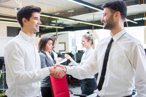 Two smiling businessmen shaking hands while standing in the office together with business people in the background.