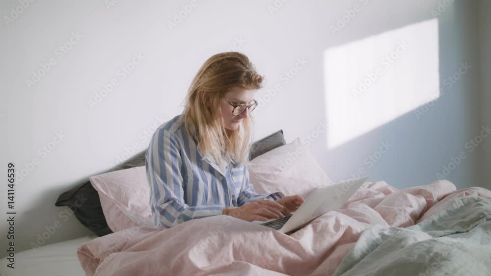 Young blonde girl in glasses wearing her pyjamas is sitting in the bed and working on her laptop from home at morning Young blonde girl in glasses wearing her pyjamas is sitting in the bed and working on her laptop from home at morning