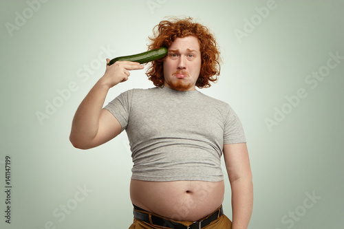 Obese overweight young redhead male ready to shoot himself out of improvised cucumber gun, having sad unhappy expression, can't stand strict vegetable diet anymore, giving up fighting against obesity