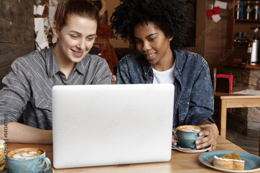 Two female students working on common university project using laptop computer together at cafe. Cheerful interracial lesbian couple browsing internet on notebook pc during lunch at restaurant Two female students working on common university project using laptop computer together at cafe. Cheerful interracial lesbian couple browsing internet on notebook pc during lunch at restaurant