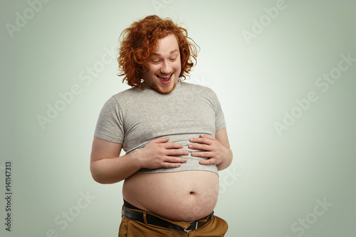 Indoor shot of funny overweight young Caucasian male wearing undersized grey t-shirt, holding hands on his stomach, looking at his big belly hanging out of pants after eating fast food on dinner