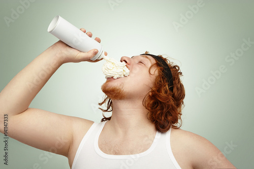 Studio shot of obese overweight Caucasian man with ginger curly hair throwing head back, spraying whipped cream in his mouth, having joyful look. People, unhealthy lifestyle, obesity and gluttony