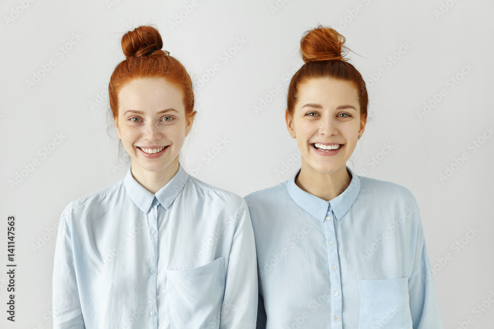 Studio shot of two Caucasian siblings with same ginger hair buns, wearing similar light-blue t-shirts, smiling happily, looking at camera, posing at white blank wall, standing to each other closely Studio shot of two Caucasian siblings with same ginger hair buns, wearing similar light-blue t-shirts, smiling happily, looking at camera, posing at white blank wall, standing to each other closely