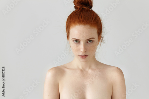 People, youth and beauty. Isolated portrait of attractive young Caucasian female with ginger hair knot posing topless indoors, having freckles all over her face and shoulders, staring at camera