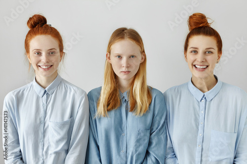 Indoor shot of three Caucasian females in similar shirts posing at blank studio wall: serious teenage girl with long loose hairstyle standing between her smiling redhead sisters with same hair buns