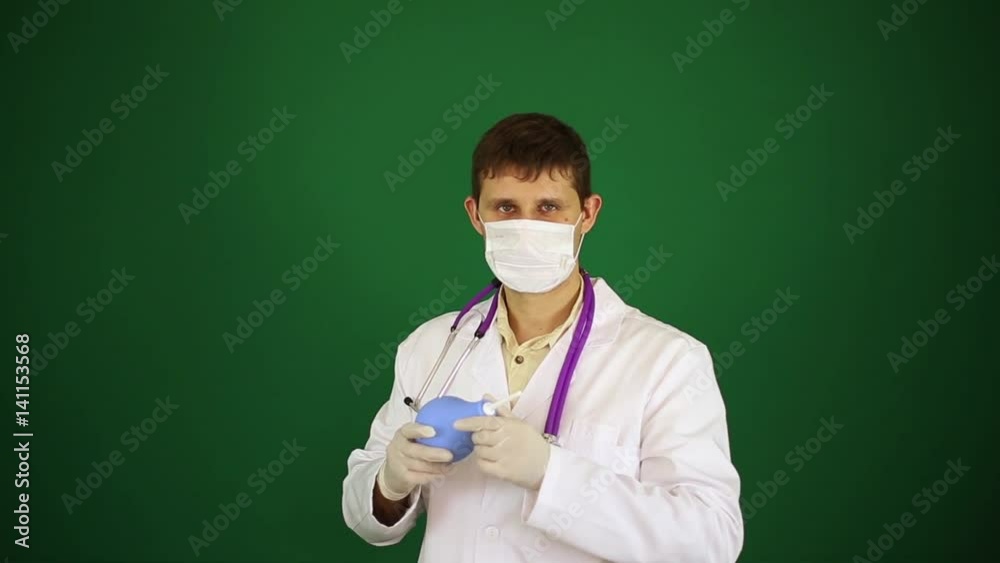 A doctor in a mask holds an enema. Doctor in white lab coat on green background. Doctor with an enema. A doctor in a mask holds an enema. Doctor in white lab coat on green background. Doctor with an enema.