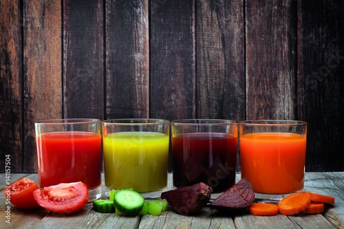 Four glasses of healthy vegetable juice with scattered vegetables and a dark wood background