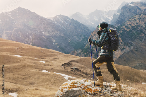 A guy with a beard and wearing sunglasses in a membrane jacket, hat, with a backpack and sticks for Nordic walking, a traveler standing and looking at the mountains