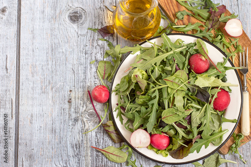  Fresh green salad with radish. Herbs such as chard, lettuce, beet leaves on a white metal plate on a cutting board, next to a glass jug with olive oil.