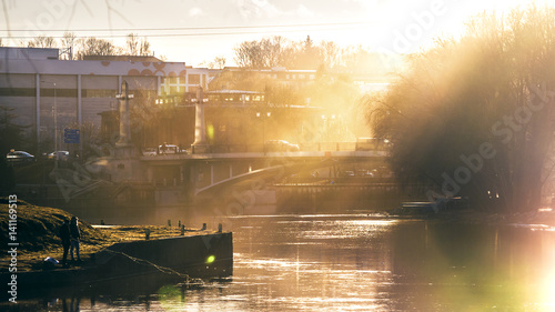 A cityscape of Tartu at evening with a river, a bridge, people fishing and prominent sun beams
