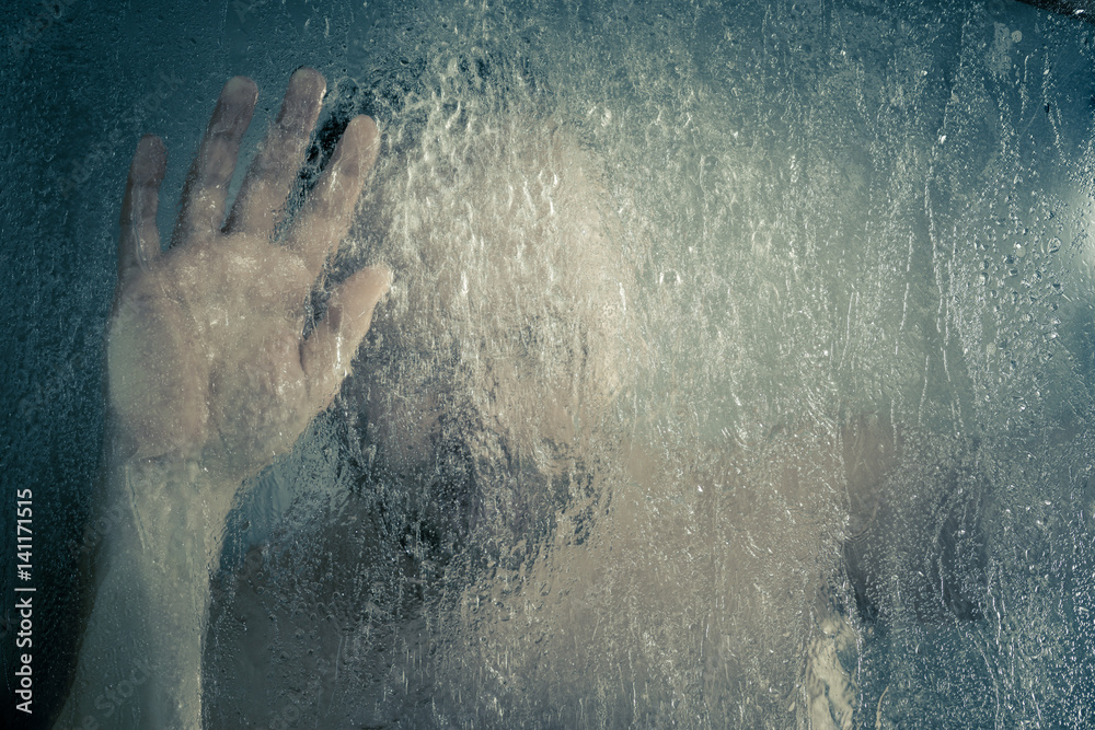 stressed man taking a shower standing under flowing water and holding his head in shower cabin behind transparent misted glass door in the bathroom