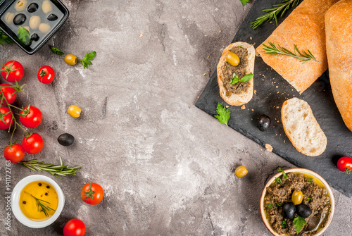 Homemade traditional Italian appetizer tapenade from green and black olives, white ciabatta, fragrant herbs and oil, fresh tomatoes. On a concrete gray table, copy space top view photo
